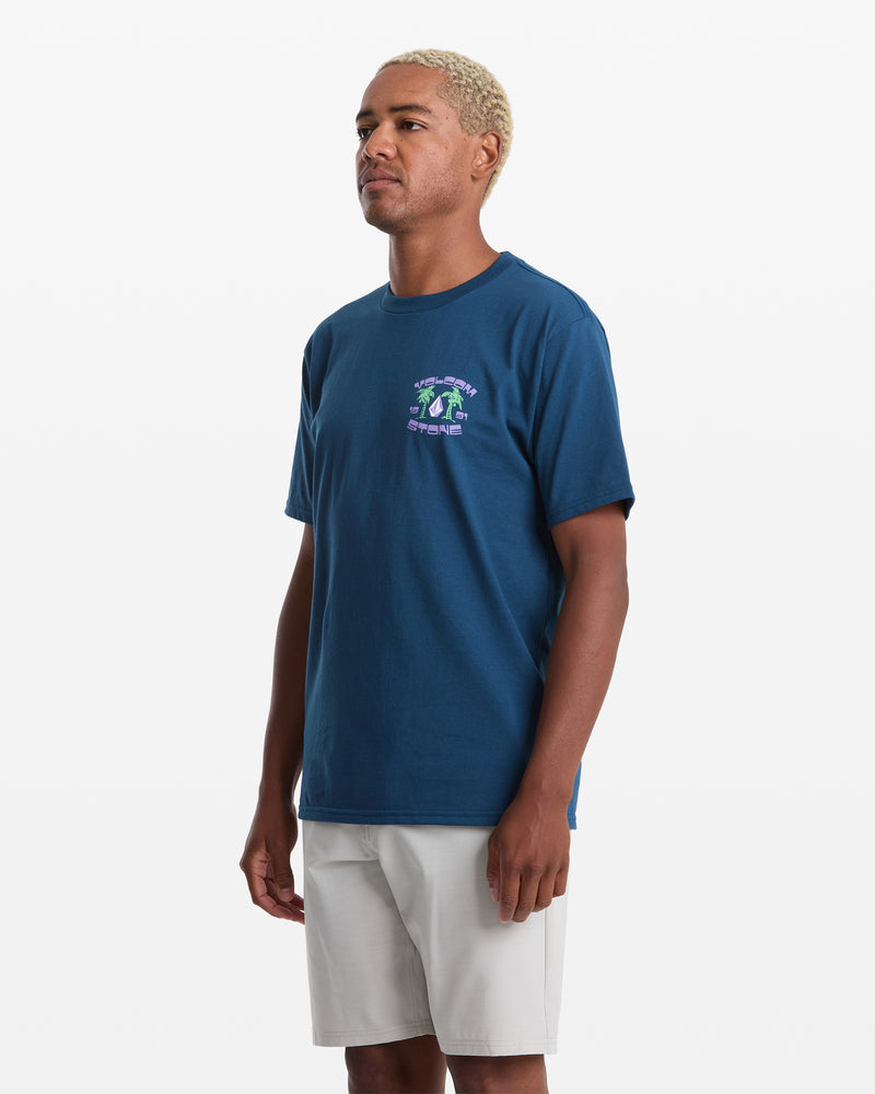 A man with short, light blond hair wears a blue Volcom Men's Fed Up Short Sleeve Tee in a classic fit and light gray shorts, standing slightly turned left against a plain white background.