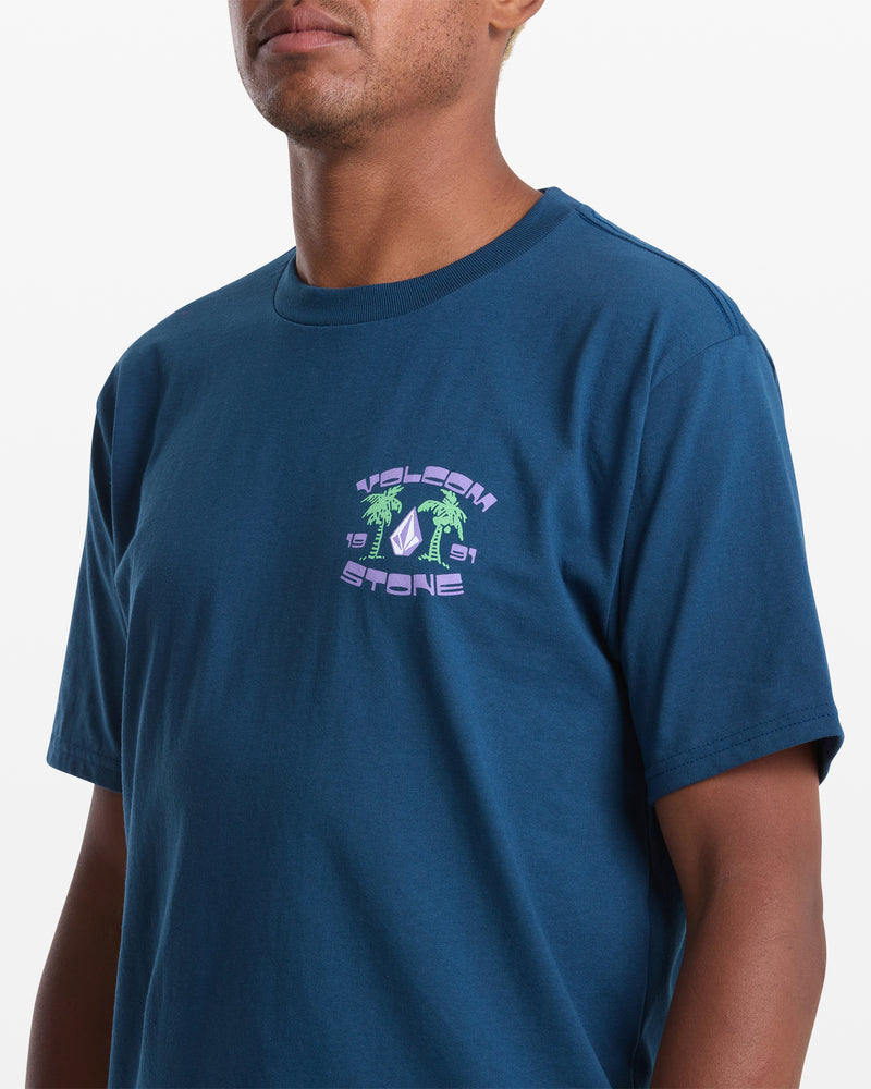 A person wears the Volcom Men's Fed Up Short Sleeve Tee, a classic fit 100% cotton blue shirt with a small chest graphic featuring palm trees, "Yaleen," and stylized purple and green text. Plain white background.