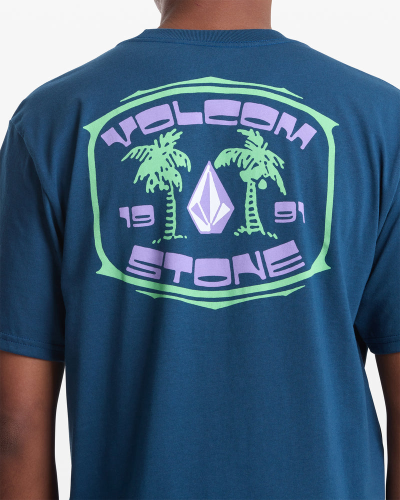 A person wearing a blue Volcom Men's Fed Up Short Sleeve Tee featuring 100% cotton, a back graphic with two palm trees, the Volcom Stone logo, and "VOLCOM 1991 STONE" text in green and purple.