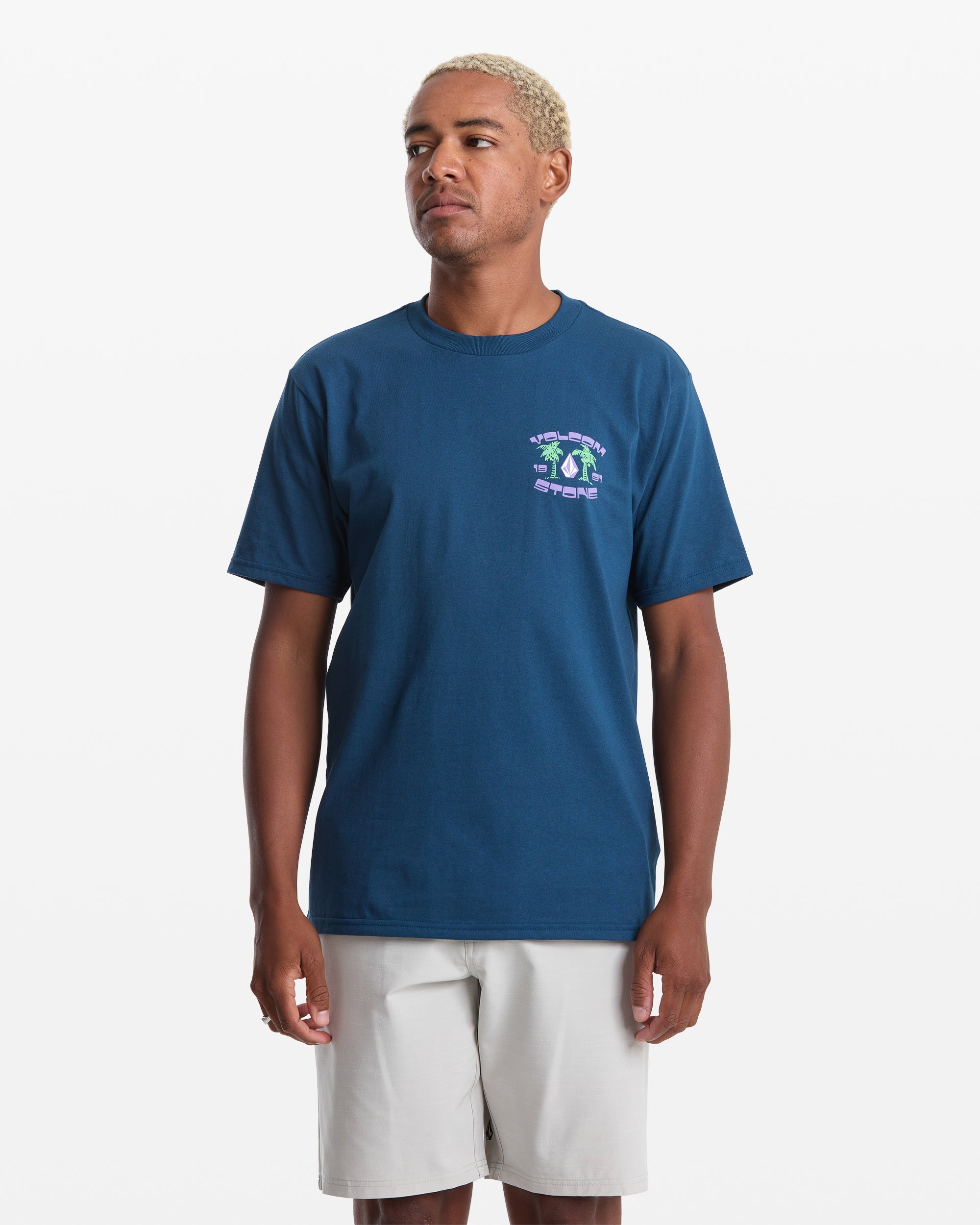 A person with short, curly blond hair wears the Volcom Men's Fed Up Short Sleeve Tee in navy blue—classic fit, 100% cotton, with a small colorful chest graphic—paired with light gray shorts, standing against a plain white background.