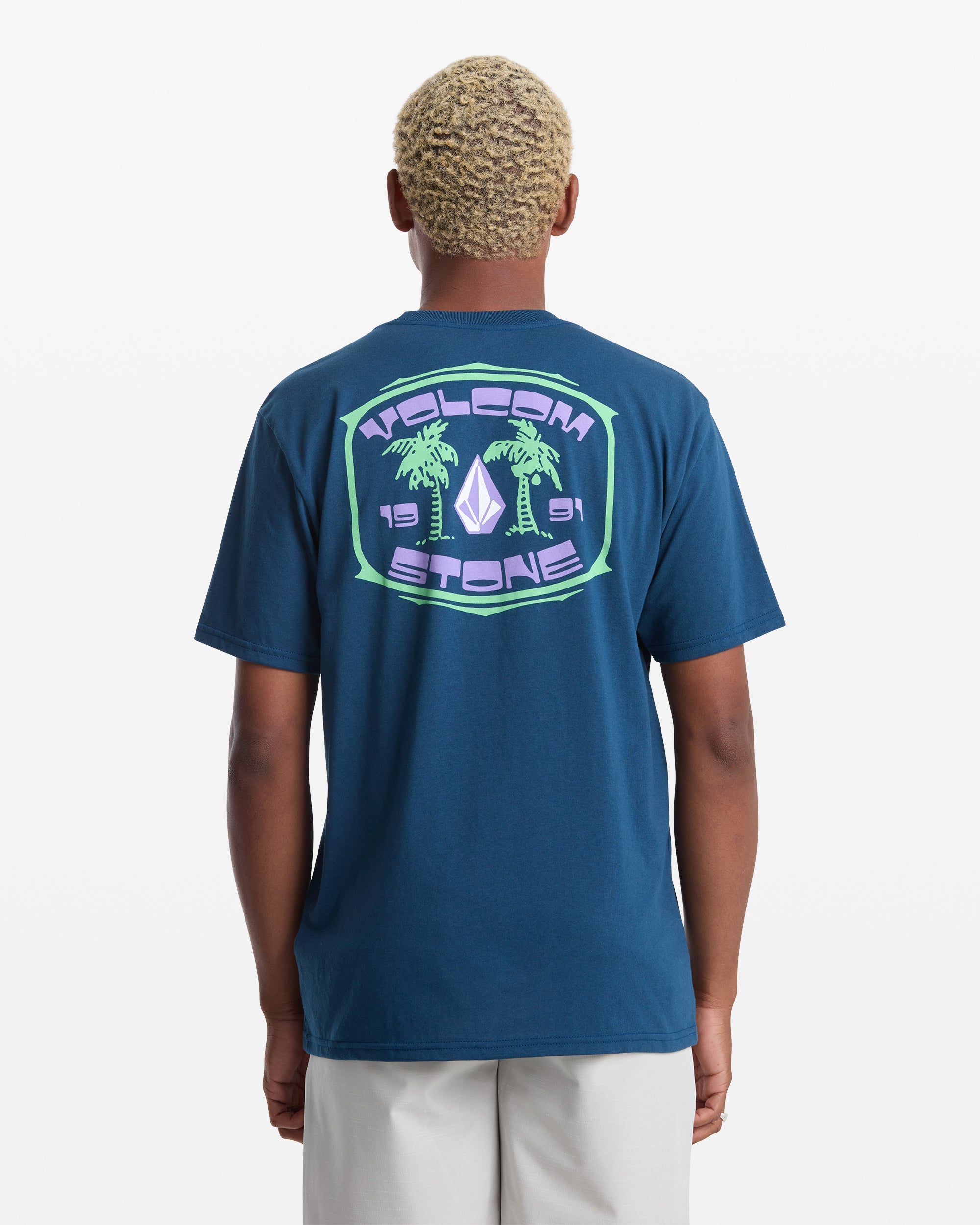 A person with short, light curly hair wears the Volcom Men's Fed Up Short Sleeve Tee, a classic fit 100% cotton blue shirt featuring a colorful back graphic with two palm trees, a diamond shape, and "VOLCOM STONE 1991" on a plain white background.