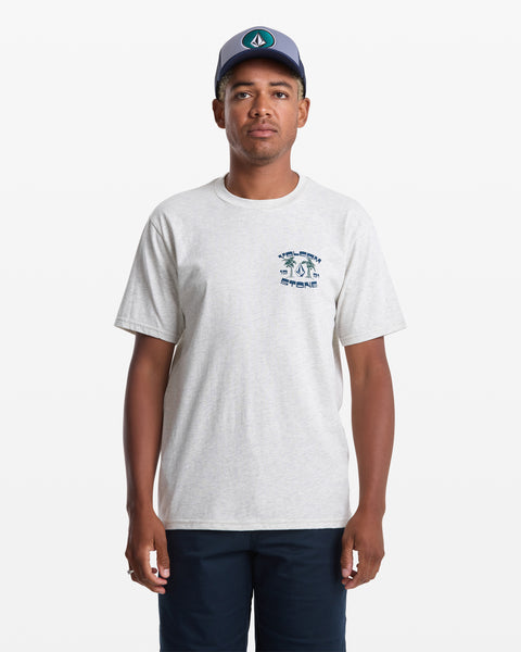 A man stands against a plain white background wearing the Volcom Men's Fed Up Short Sleeve Tee in light gray with a small soft-hand screen print on the left chest, paired with navy shorts and a blue baseball cap.