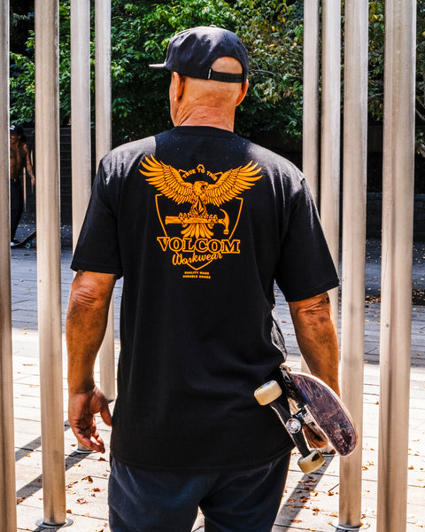 A man in a Volcom Men's Hammered Split Short Sleeve Tee—black, 100% cotton with a yellow eagle graphic—stands outdoors holding a skateboard, facing away toward tall metal poles.