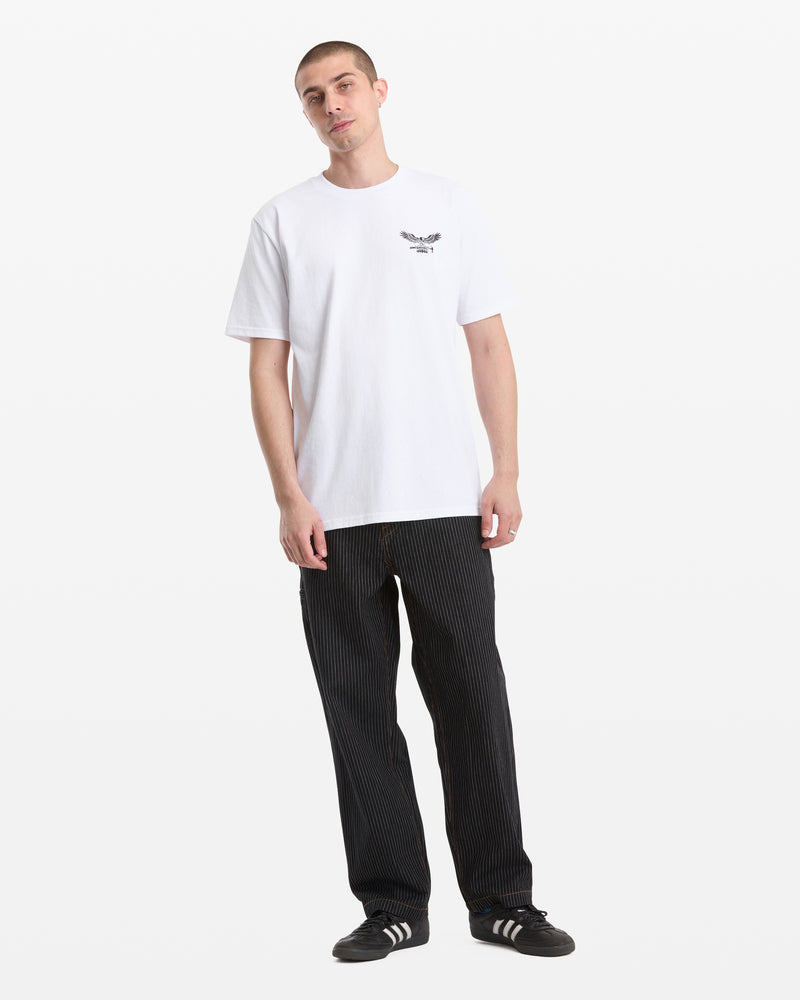 A man faces forward in a Volcom Men's Hammered Split Short Sleeve Tee, featuring a small black chest graphic, paired with loose black pinstripe pants and black Adidas sneakers with white stripes. The background is plain white.