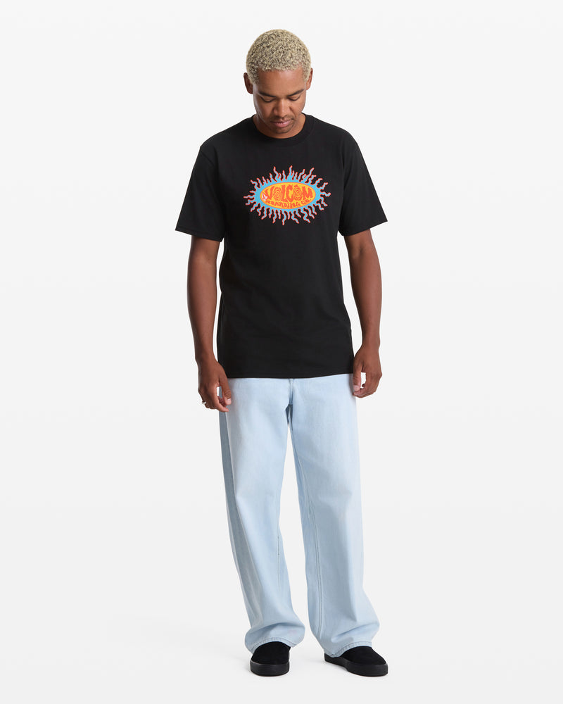A person with short, bleached hair wears the Volcom Men's Live Wire Short Sleeve Tee, featuring a colorful sunburst-style logo, paired with light blue jeans and black shoes, standing against a plain white background.