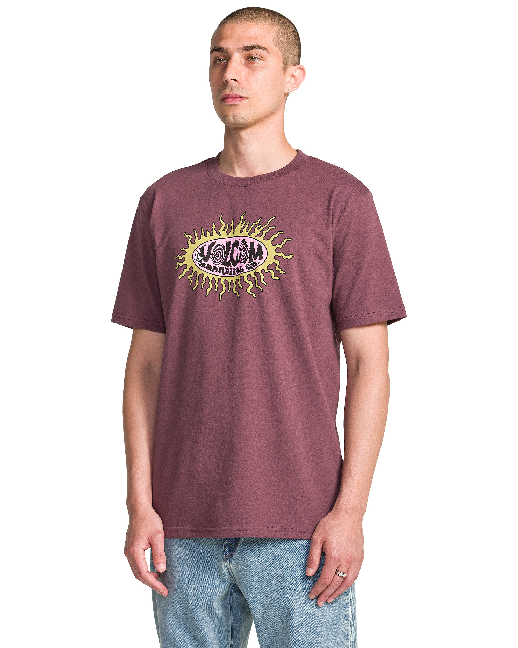 A man with short hair wears the Volcom Men's Live Wire Short Sleeve Tee, a maroon shirt with a circular yellow sun-style graphic and "VOLCOM" at the center, paired with light blue jeans, standing before a plain white backdrop.