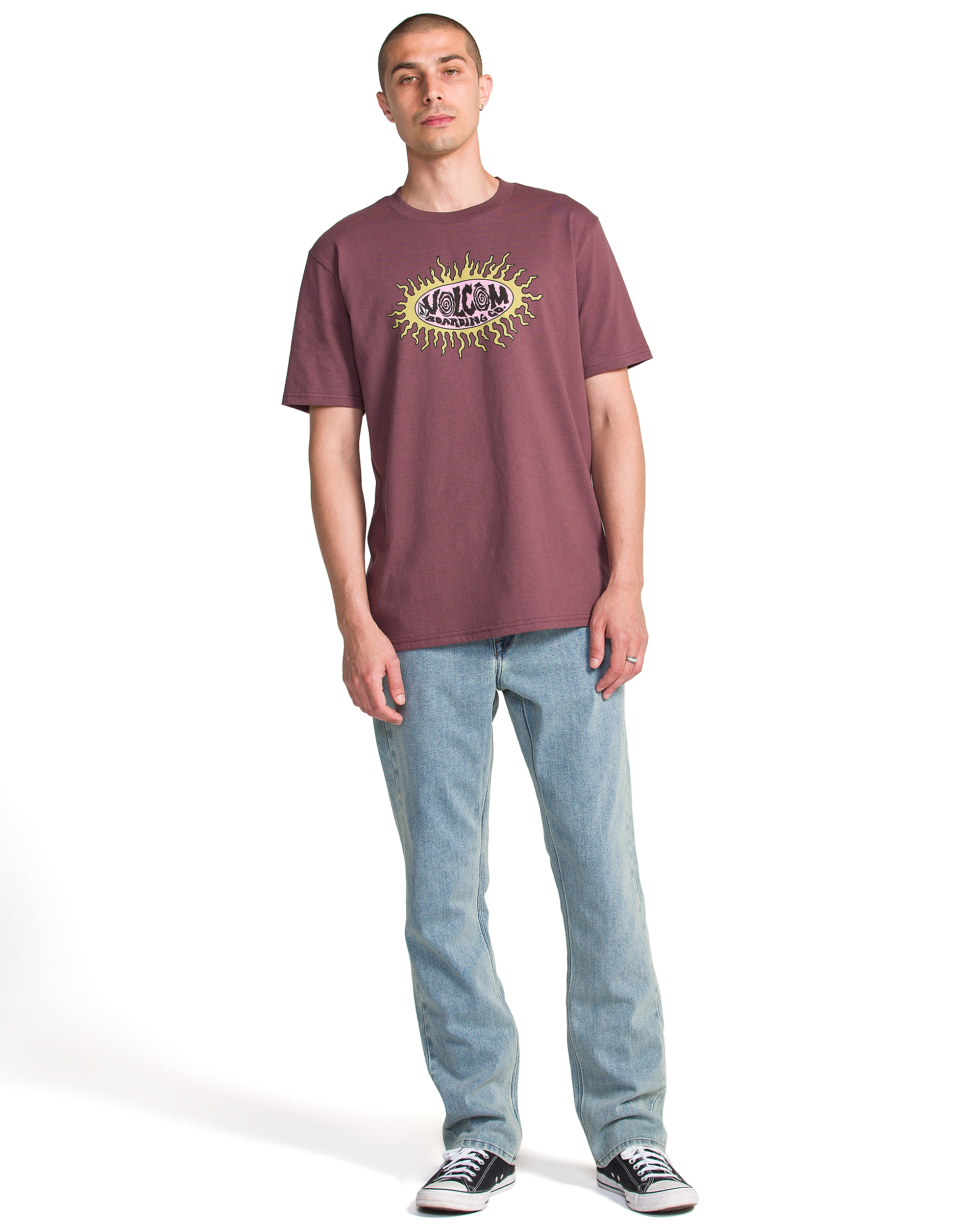 A man stands against a white background wearing the Volcom Men's Live Wire Short Sleeve Tee in maroon, light blue jeans, and black sneakers.