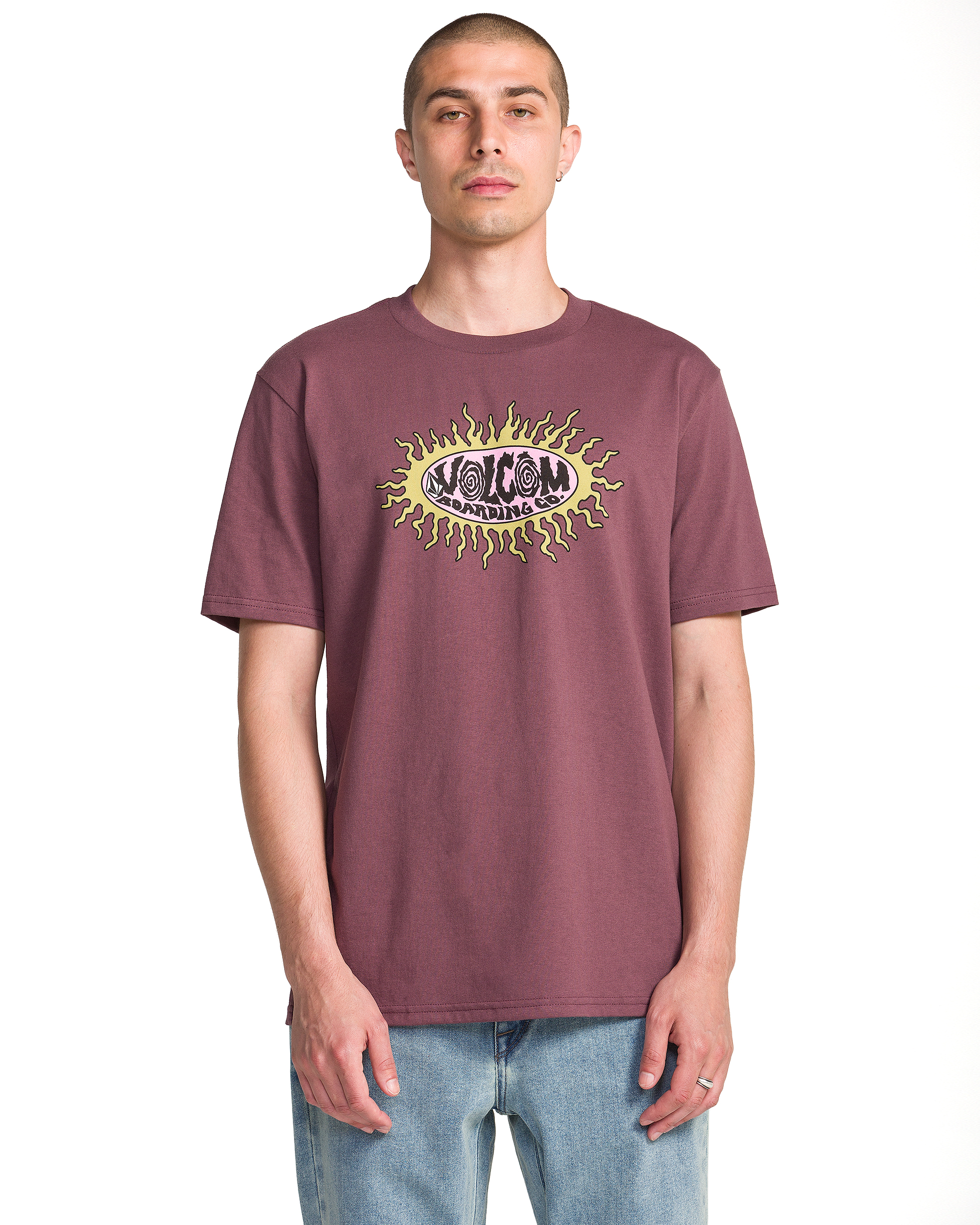 A person with short hair wears the Volcom Men's Live Wire Short Sleeve Tee—a maroon shirt with a bold sun-ray logo and VOLCOM STONE text—paired with light blue jeans, standing against a white background.