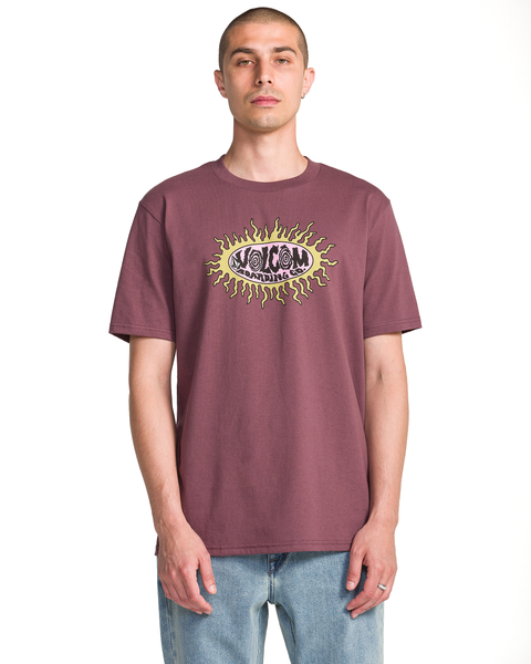 A person with short hair wears the Volcom Men's Live Wire Short Sleeve Tee—a maroon shirt with a bold sun-ray logo and VOLCOM STONE text—paired with light blue jeans, standing against a white background.