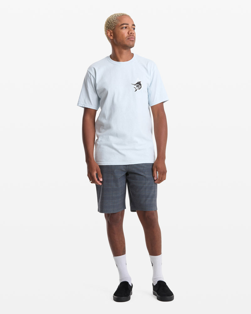 A person with short blond hair wears the Volcom Men's Marlinsane Short Sleeve Tee, gray plaid shorts, white socks, and black slip-on shoes while standing against a plain white background.