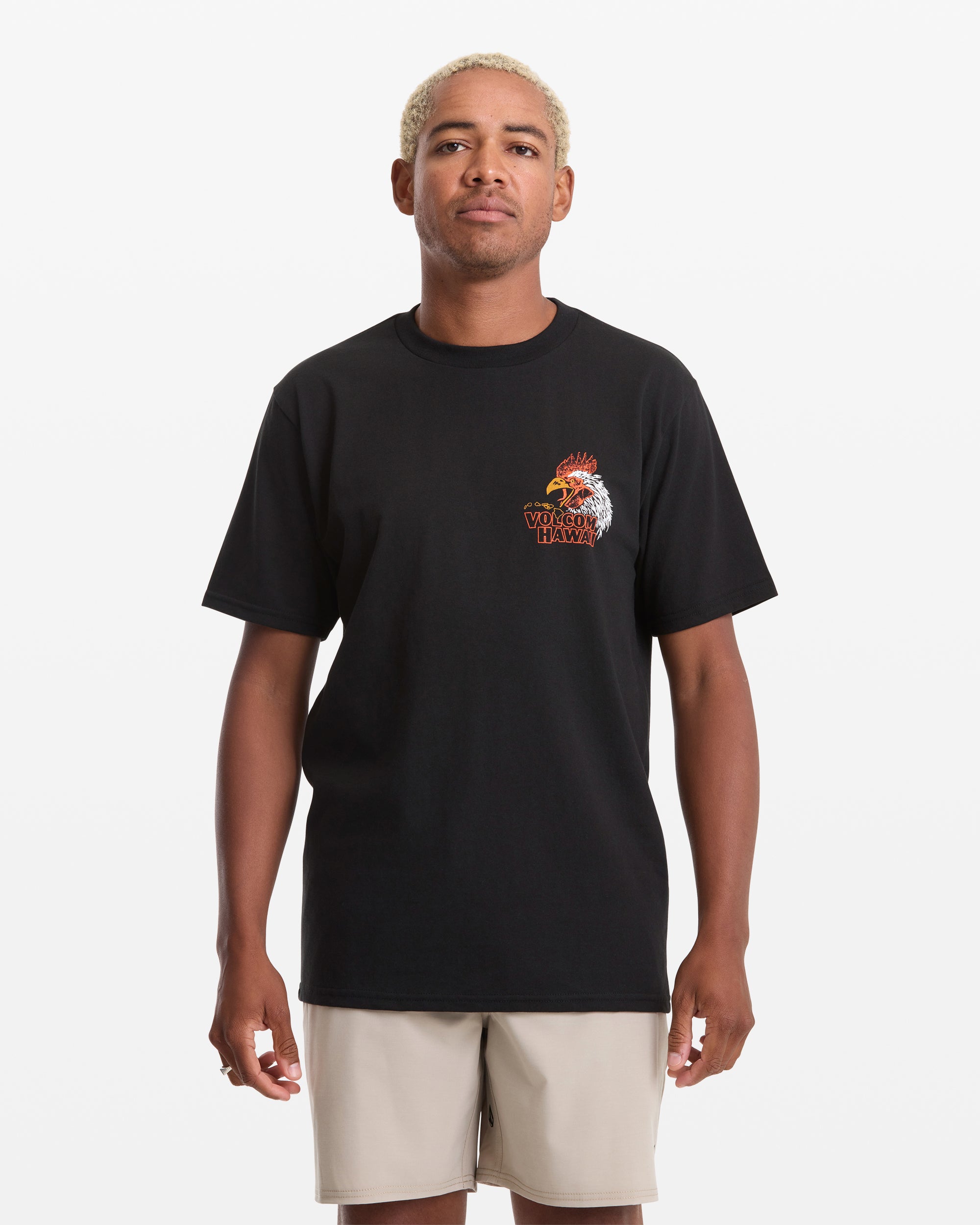 A person with short blond hair wears the Volcom Men's Yewster Rooster Short Sleeve Tee—a black T-shirt with a rooster graphic and text on the left chest—paired with light shorts, standing against a white background.