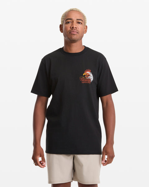 A person with short blond hair wears the Volcom Men's Yewster Rooster Short Sleeve Tee—a black T-shirt with a rooster graphic and text on the left chest—paired with light shorts, standing against a white background.