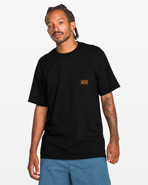 A man stands against a plain white background wearing the Volcom Men’s Workwear Certifico Short Sleeve Tee—a classic fit black T-shirt made from premium cotton with a small orange woven label patch on the pocket—paired with blue shorts.
