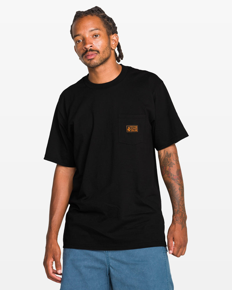 A man stands against a plain white background wearing the Volcom Men’s Workwear Certifico Short Sleeve Tee—a classic fit black T-shirt made from premium cotton with a small orange woven label patch on the pocket—paired with blue shorts.