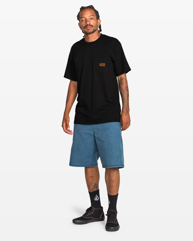 A man with short, braided hair and tattoos on one arm stands against a white background, wearing the Volcom Men’s Workwear Certifico Short Sleeve Tee in classic black, paired with blue knee-length shorts, black socks featuring a white logo, and black sneakers.