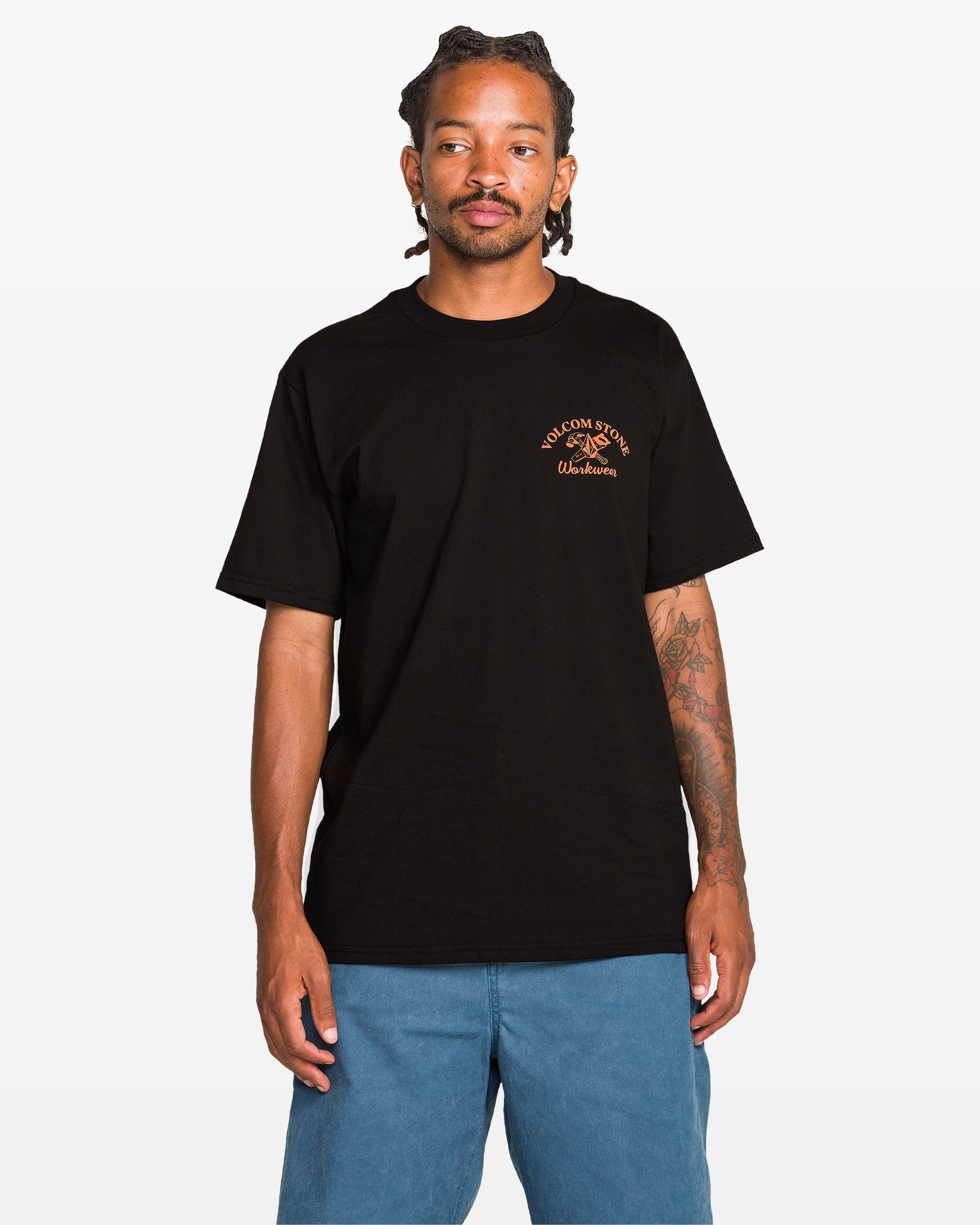 A man with dreadlocks and tattoos on his left arm models the Volcom Men's Crosscut Workwear Short Sleeve Tee in black, featuring a small orange chest logo, paired with blue shorts against a plain white background.