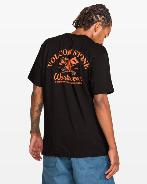 A man with dreadlocks faces away, wearing a Volcom Men's Crosscut Workwear Short Sleeve Tee in classic fit with bold orange graphics on the back, paired with blue all-cotton shorts.