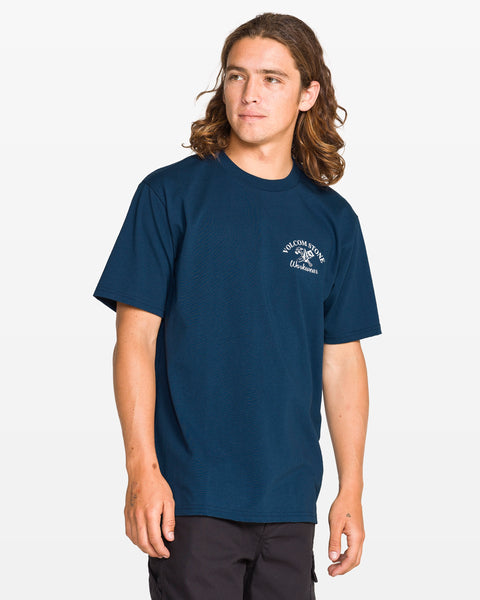 A person with long brown hair wears a dark blue Volcom Men's Crosscut Workwear Short Sleeve Tee and black pants, standing against a white background. The all-cotton shirt has a small white logo and text on the left chest.