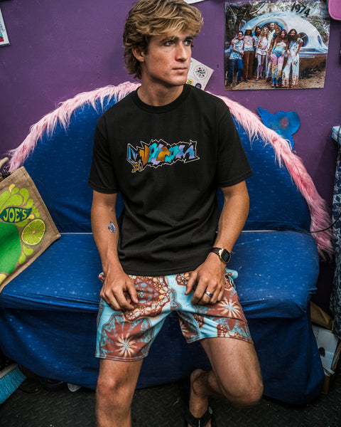 A young man sits on a blue couch with pink fuzzy trim, wearing the Volcom Men's Star Shields Featured Artist Exp Short Sleeve Tee and colorful shorts. Behind him, a purple wall displays photos and Star Shields artwork.