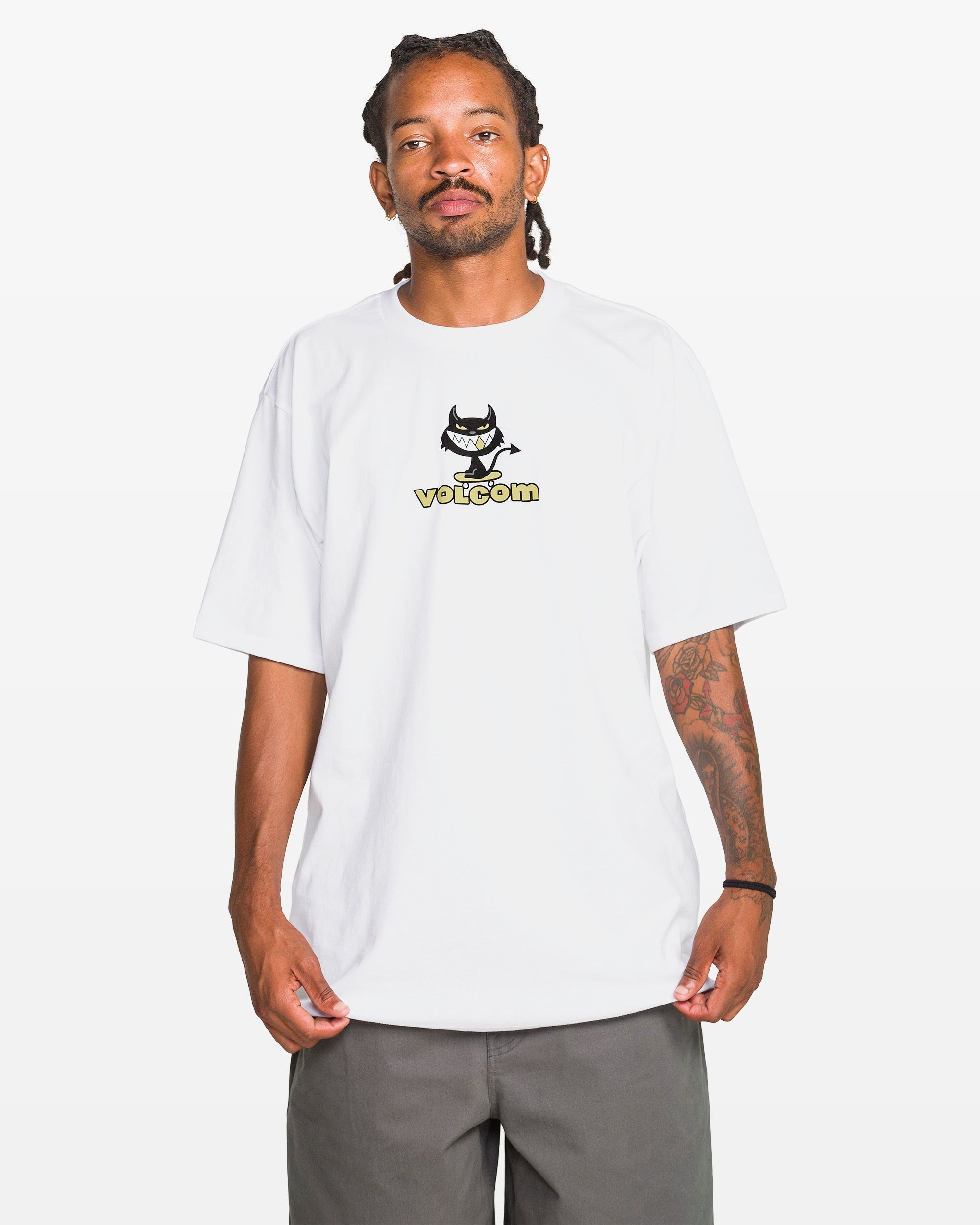A person wears the Volcom Men's Devilish Short Sleeve Tee, a classic fit white 100% cotton T-shirt with a small cartoon devil graphic and Volcom text, standing against a plain white background.