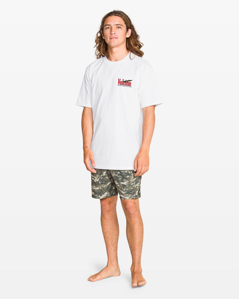 A person with long hair stands barefoot on a white background, wearing the Volcom Men's Stoney Mountain Short Sleeve Tee, a classic fit 100% cotton tee with a red and black chest logo, paired with camouflage shorts.