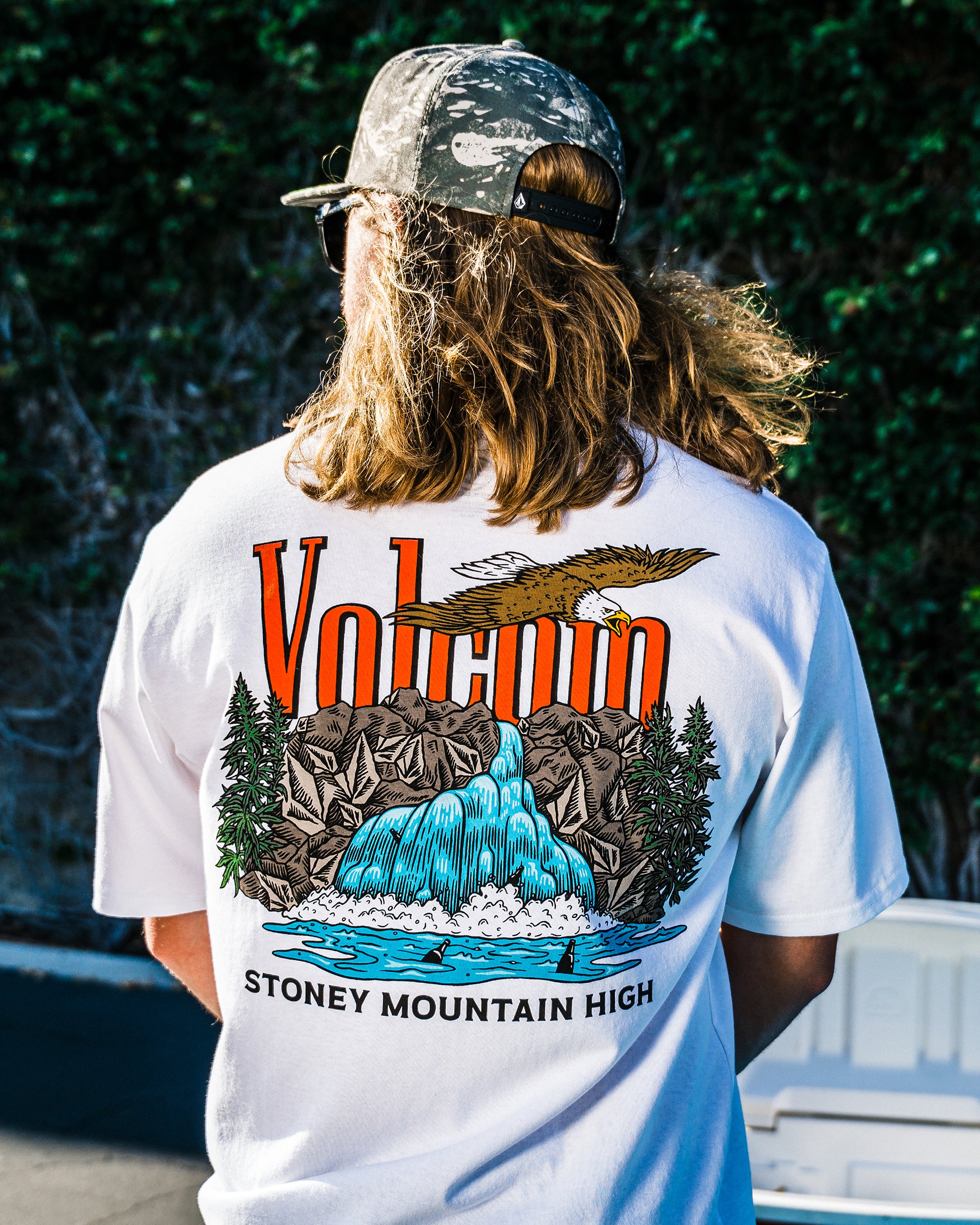 A person with long hair wears a camouflage cap and the Volcom Men's Stoney Mountain Short Sleeve Tee, made from 100% cotton with a vibrant waterfall, mountains, eagle, and trees graphic, plus VOLCANO and STONEY MOUNTAIN HIGH on the back.