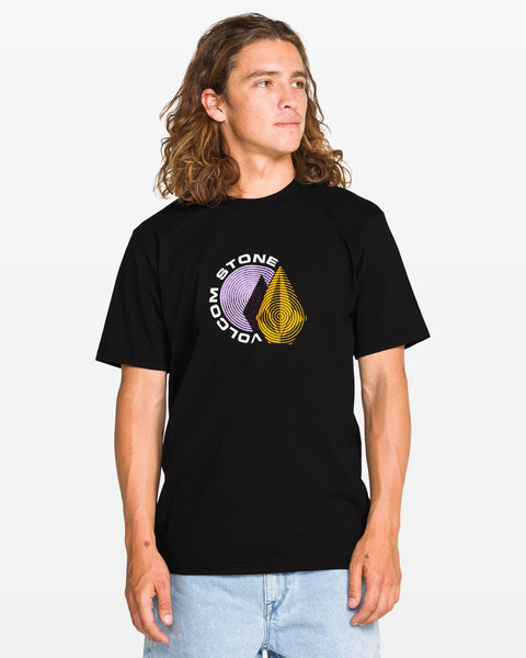 A young man with long wavy hair wears a black T-shirt with a circular Volcom Stone logo in purple and yellow on the chest. He stands facing slightly to the side, hands relaxed, against a plain white background.