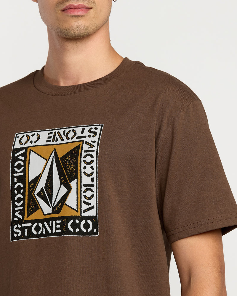 A man wears the Volcom Men's Angle Iron Short Sleeve Tee in brown, featuring a geometric logo and "VOLCOM STONE CO." on the chest. Only his lower face and upper torso show. Made from premium ring-spun cotton for comfort.