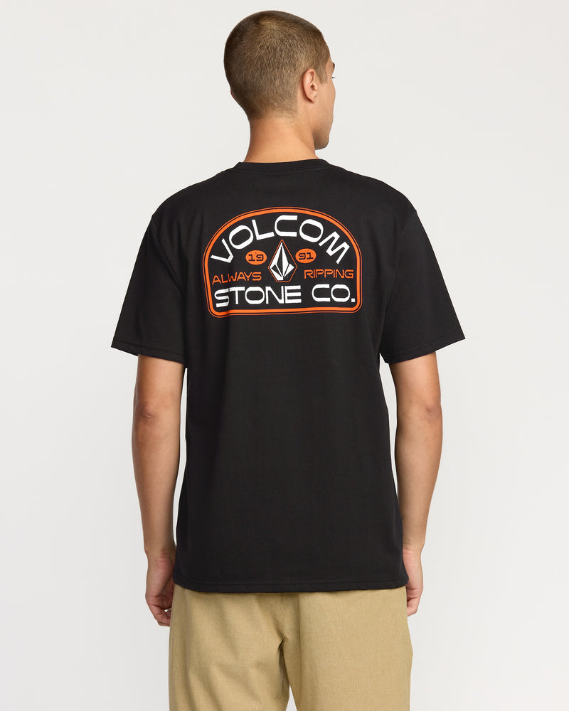 A person wears the Volcom Men's Always Ripping Short Sleeve Tee in black, featuring a classic fit and large back logo, made from premium ring spun cotton, paired with tan pants against a plain white background.