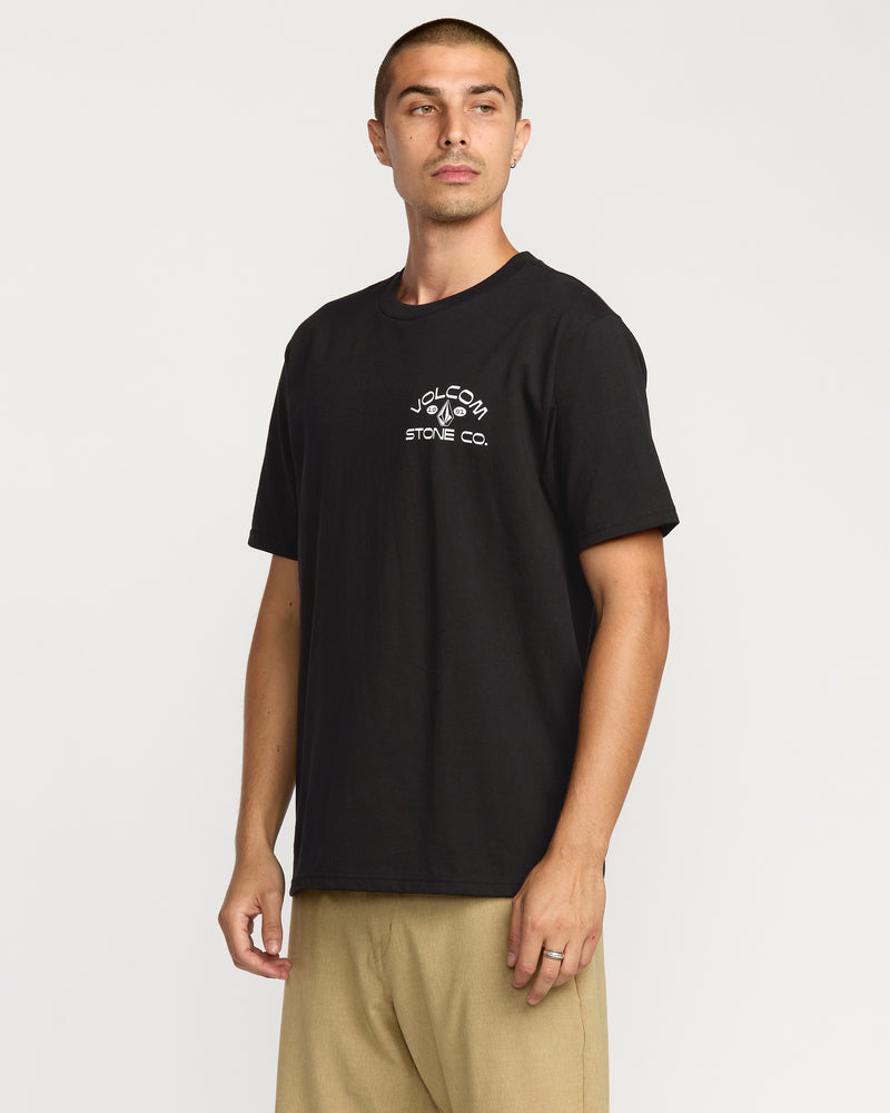 A man with short hair wears a black Volcom Men's Always Ripping Short Sleeve Tee in a classic fit and beige pants, standing against a plain white background and looking slightly to his left.