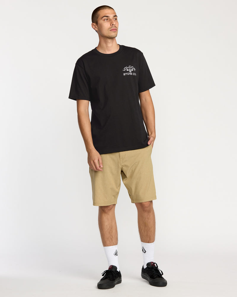 A man stands against a white background wearing the Volcom Men's Always Ripping Short Sleeve Tee in black, tan shorts, white crew socks, and black sneakers. His left hand is in his pocket and he looks slightly to the left.