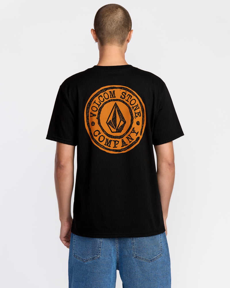 A person with a shaved head, dressed in the Volcom Men’s Logo Stamp Short Sleeve Tee—crafted from premium ring spun cotton and featuring an orange circular logo on the back—paired with blue jeans, stands with their back to the camera against a plain white background.