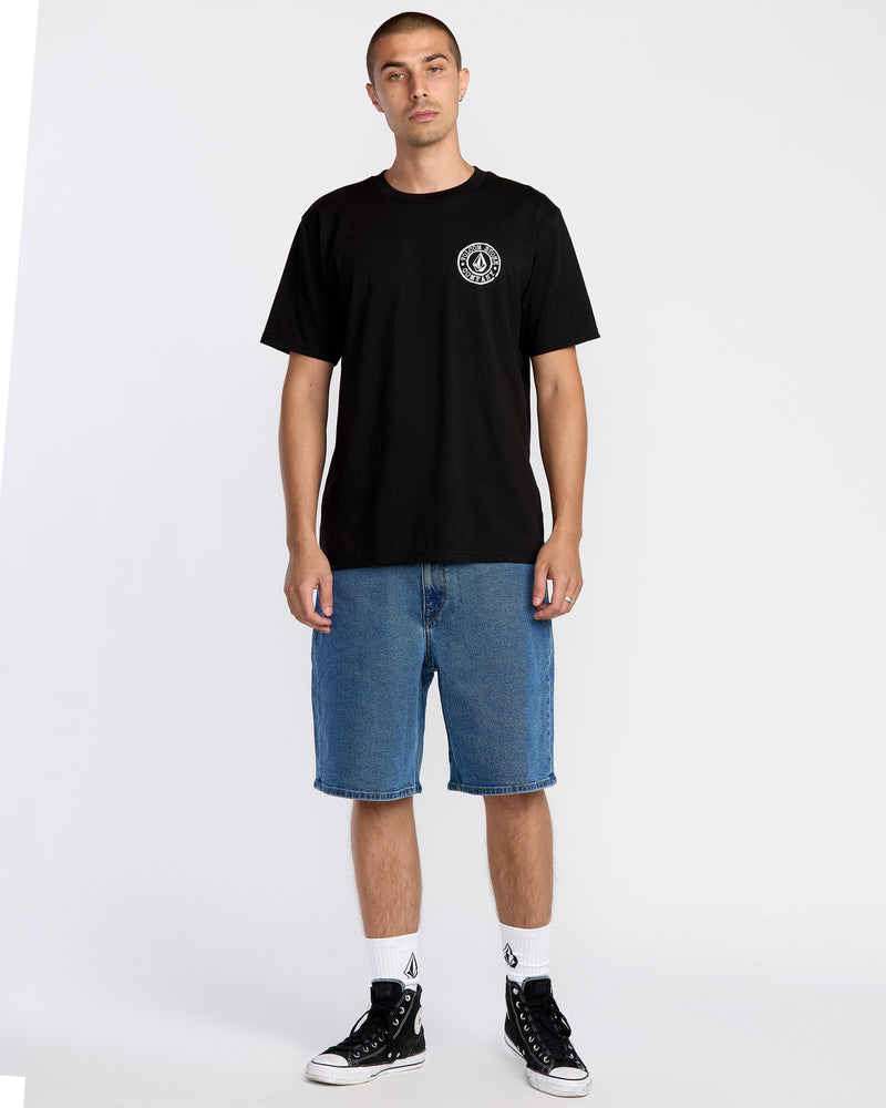 A person stands against a plain background wearing the Volcom Men’s Logo Stamp Short Sleeve Tee, crafted from premium ring spun cotton with a small white logo, paired with blue denim shorts, white socks featuring a black logo, and black sneakers.
