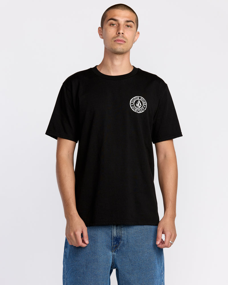 A person wearing the Volcom Men’s Logo Stamp Short Sleeve Tee in black, featuring a small white circular logo on the chest, and blue jeans stands against a plain white background.