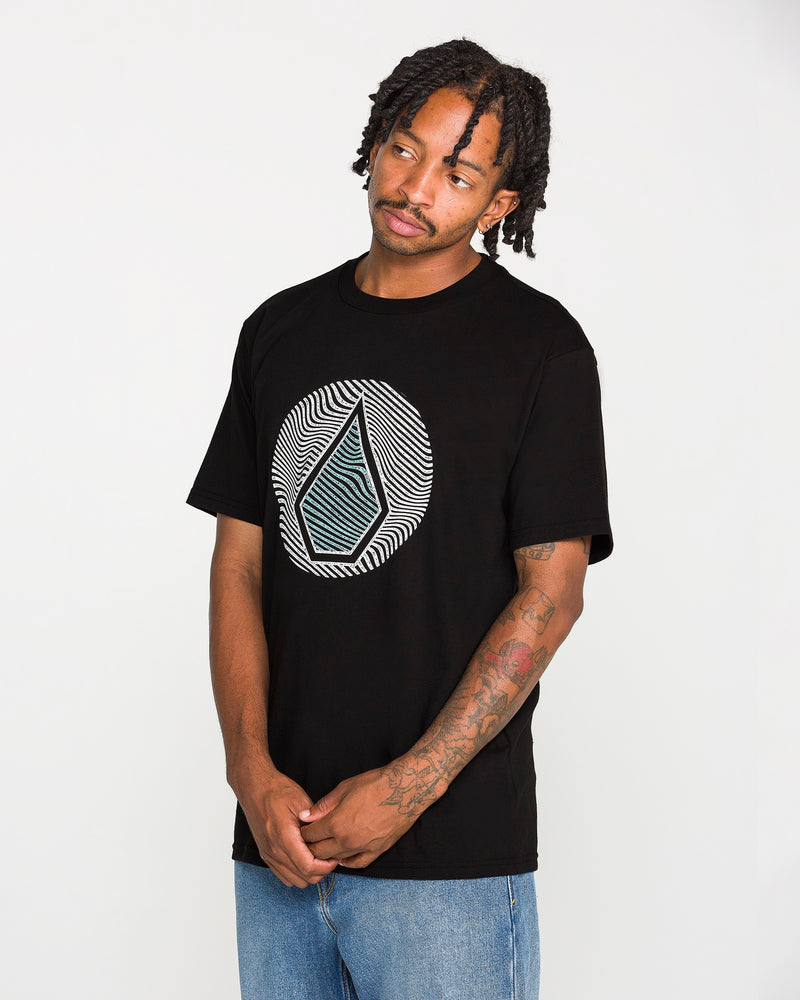A man with braided hair, a mustache, and tattoos on his forearm wears the Volcom Men's Wave Stone Short Sleeve Tee—a classic fit t-shirt made from premium ring spun cotton and featuring a geometric design. He pairs it with blue jeans and stands against a plain white background.