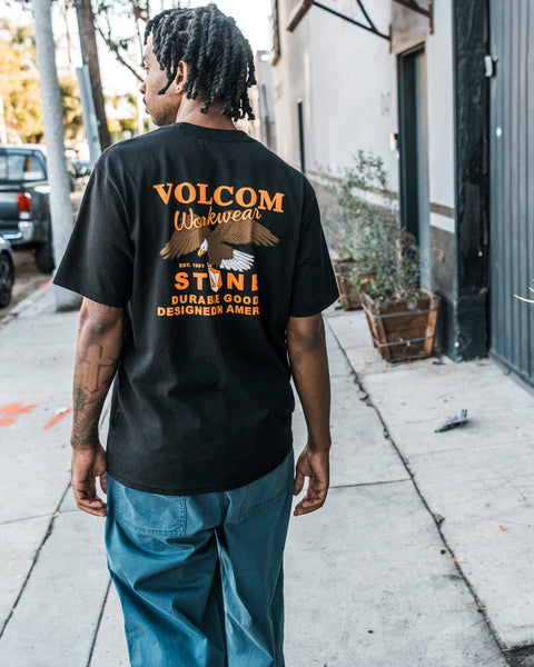 A person walks on a sidewalk lined with shrubs and buildings, wearing Volcom’s Men’s Stone Talon Workwear Short Sleeve Tee in classic fit black heavyweight cotton. The back of the shirt features orange and white “VOLCOM Workwear” text, and it’s paired with blue pants.