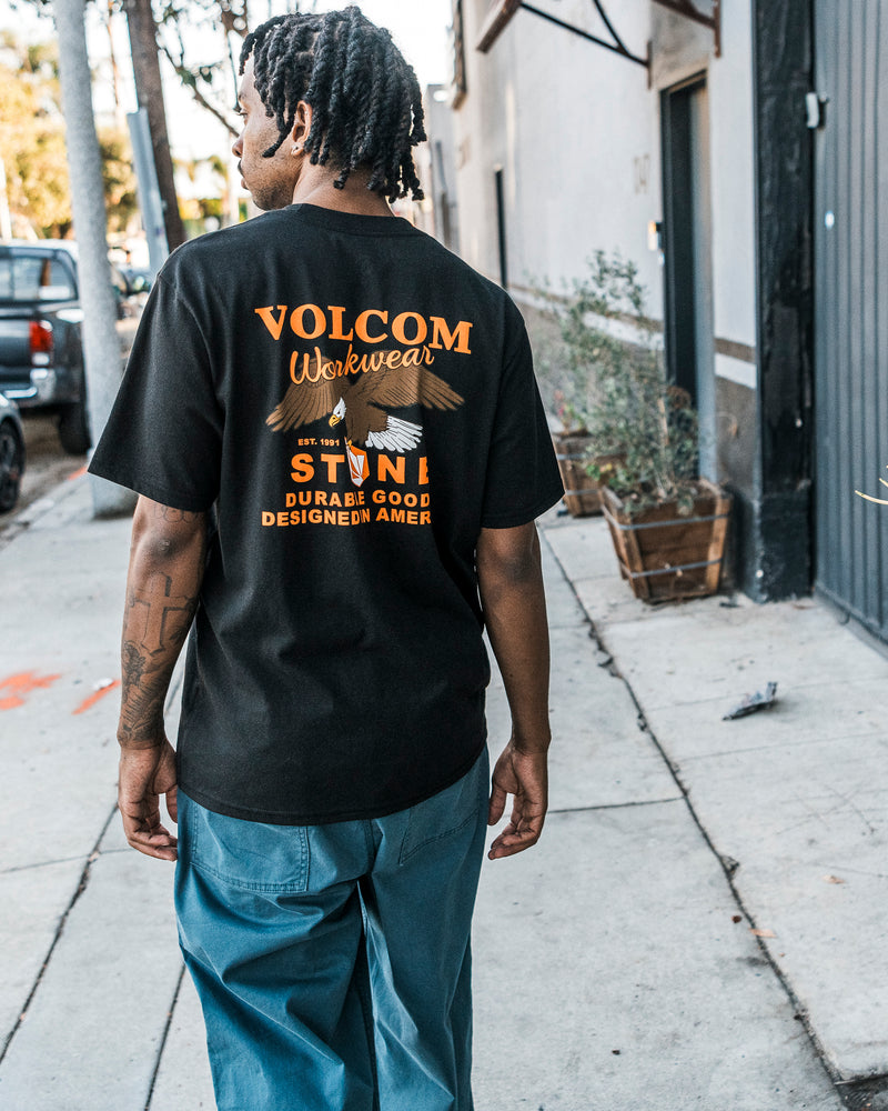 A person walks on a sidewalk lined with shrubs and buildings, wearing Volcom’s Men’s Stone Talon Workwear Short Sleeve Tee in classic fit black heavyweight cotton. The back of the shirt features orange and white “VOLCOM Workwear” text, and it’s paired with blue pants.