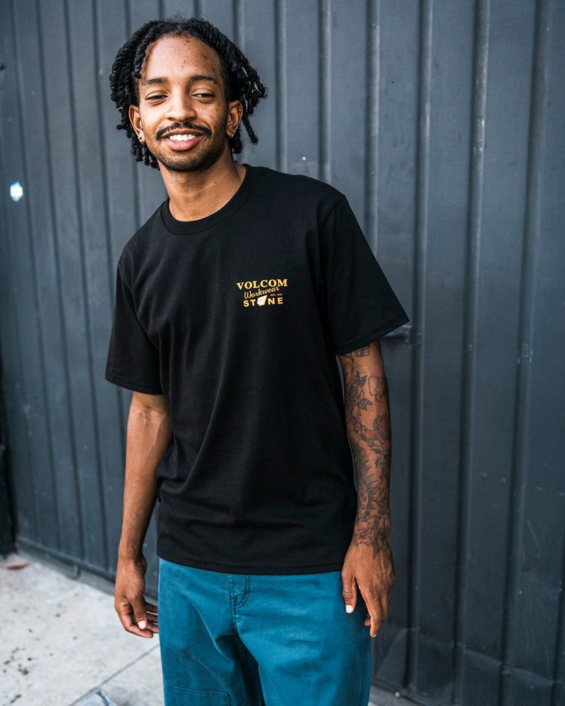 A man with short dreadlocks, dressed in blue pants and a black Volcom Men’s Stone Talon Workwear Short Sleeve Tee made from heavyweight cotton, stands smiling in front of a dark metal wall.