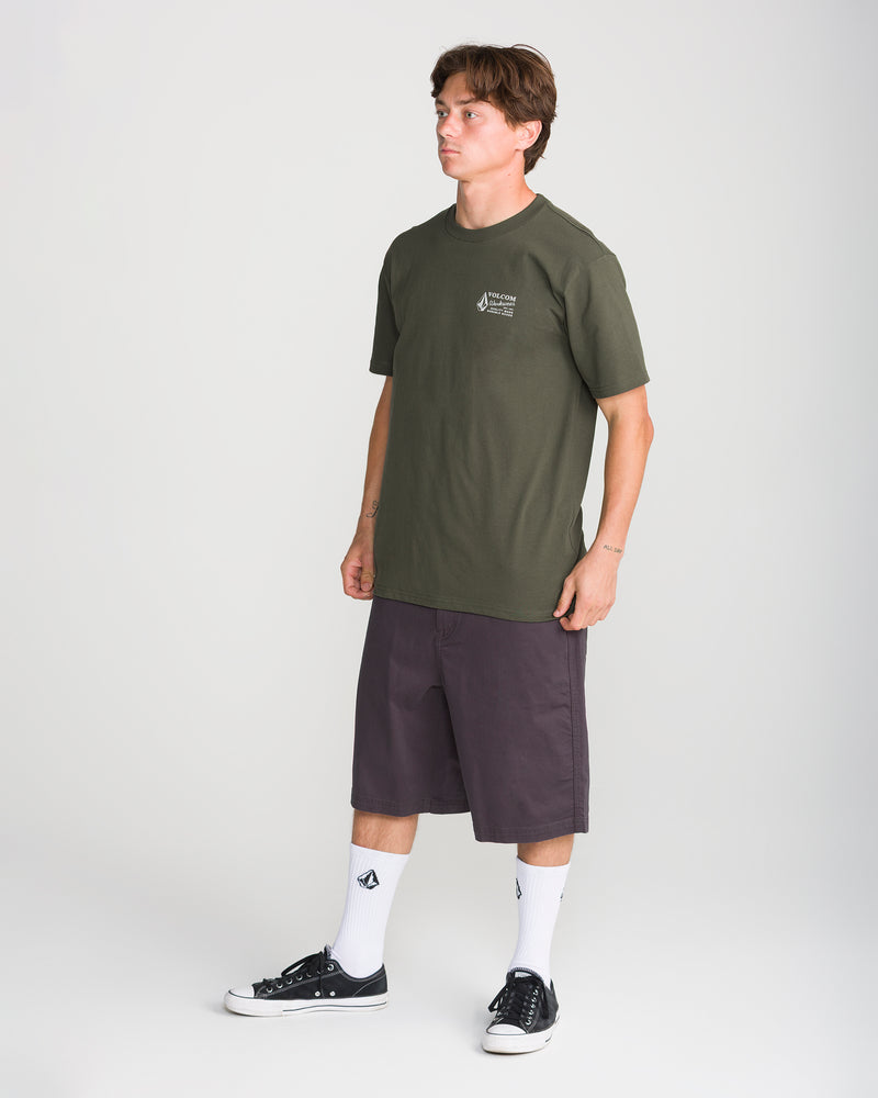 A man stands against a plain background, wearing the Men’s Volcom Workwear Short Sleeve Tee by Volcom in olive, paired with dark knee-length shorts, logo white socks, and black sneakers. He is looking to the side with his arms relaxed by his sides.