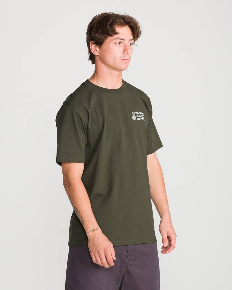 A man stands against a plain white background, looking slightly to the side, dressed in the Men’s Volcom Workwear Short Sleeve Tee. This dark green shirt features a small white Volcom logo on the chest and is constructed from heavyweight cotton for long-lasting comfort and durability.