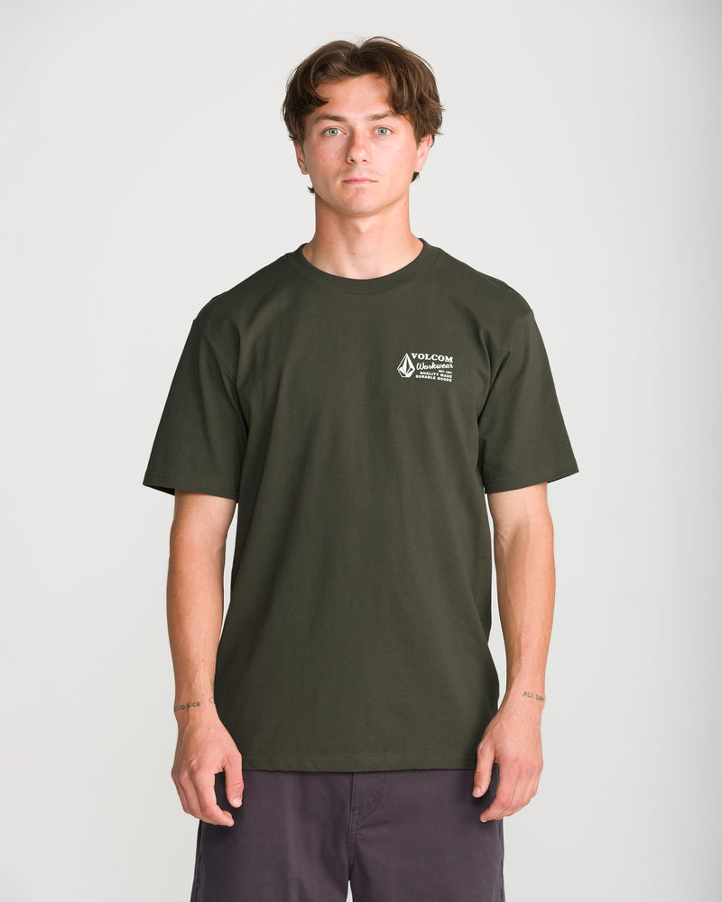 A young man stands facing forward against a plain light background, wearing the Men’s Volcom Workwear Short Sleeve Tee by Volcom in dark green heavyweight premium cotton, featuring a small white logo on the left chest, and paired with dark shorts.
