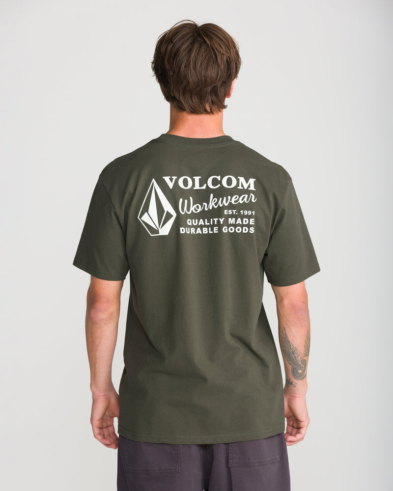 A person with short brown hair is seen from behind wearing the Men’s Volcom Workwear Short Sleeve Tee by Volcom in olive green, crafted from heavyweight premium cotton. The back of the shirt features white text and the Volcom logo, highlighting its durable goods heritage. The person pairs the tee with dark pants.