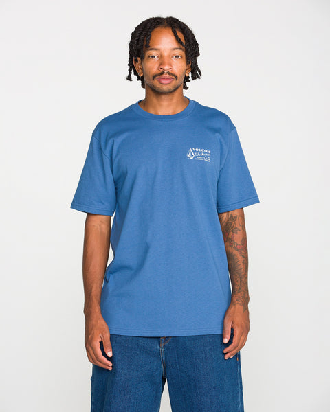 A young Black man with braided hair and a mustache stands against a white background, wearing the Volcom Men's Volcom Workwear Short Sleeve Tee in blue paired with blue jeans. He has visible tattoos on his left arm and is looking directly at the camera.