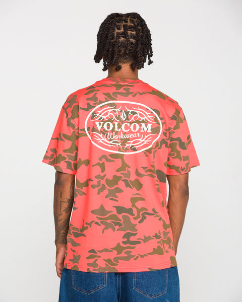 A person wearing the Volcom Men's Safety Workwear Short Sleeve Tee in pink and brown camo, crafted from premium cotton, stands with their back to the camera, highlighting the bold white logo—combining distinctive style with practical safety workwear.