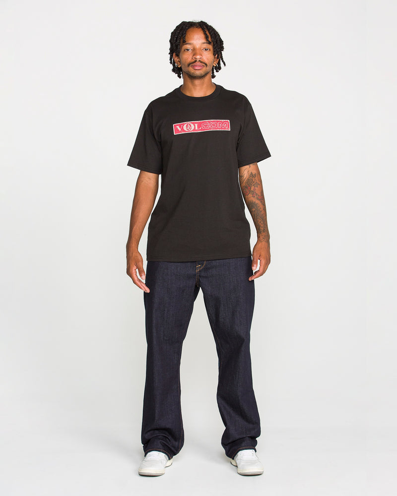 A man stands facing forward, wearing the Volcom Men's Dual Euro Short Sleeve Tee in black, paired with dark baggy jeans and white sneakers; he has a neutral expression and his arms rest relaxed by his sides against a plain white background.
