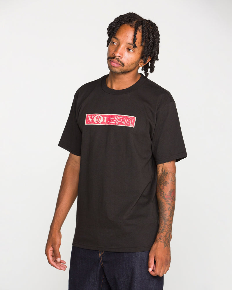 A man with short dreadlocks wears the Volcom Men's Dual Euro Short Sleeve Tee, a classic fit t-shirt made from premium ring-spun cotton and featuring a bold red and white vintage Volcom logo. He pairs it with dark jeans and stands against a plain light background, gazing slightly to the side with a relaxed expression.