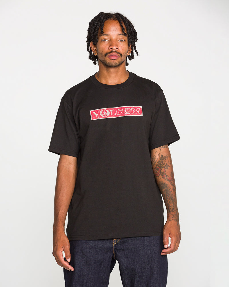 A man with tattoos stands facing the camera, wearing the Volcom Men's Dual Euro Short Sleeve Tee—a classic fit made from premium ring-spun cotton with a bold red and white vintage Volcom logo—paired with dark jeans against a plain white background.