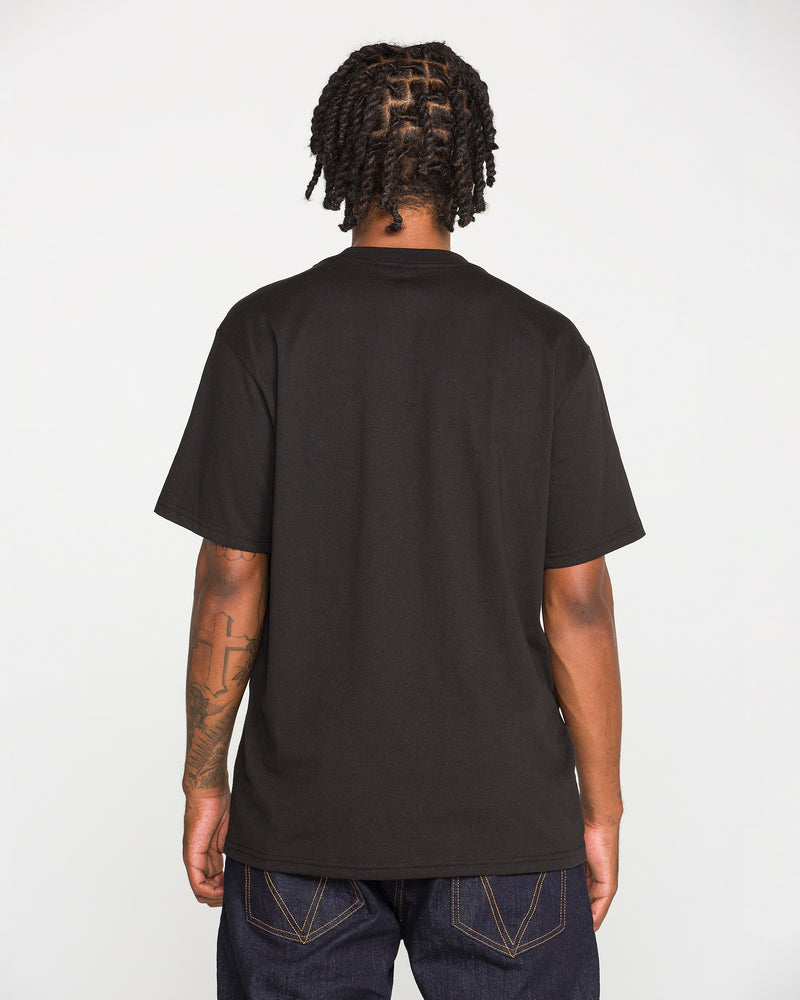 A person with braided hair, wearing the Volcom Men's Dual Euro Short Sleeve Tee in plain black and dark jeans, is standing with their back to the camera. Their arms are relaxed at their sides, revealing tattoos on one arm.