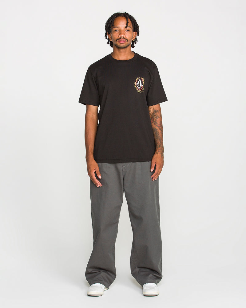 A person stands facing forward against a plain white background, wearing the Volcom Men's Ringafire Short Sleeve Tee in black—crafted from heavy-weight premium cotton with a small chest graphic—paired with gray wide-leg pants and white sneakers.