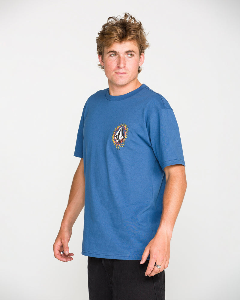 A person with tousled hair, wearing the Volcom Men's Ringafire Short Sleeve Tee in blue—crafted from premium ring spun cotton and featuring a colorful chest logo—stands turned slightly to the side against a plain white background.